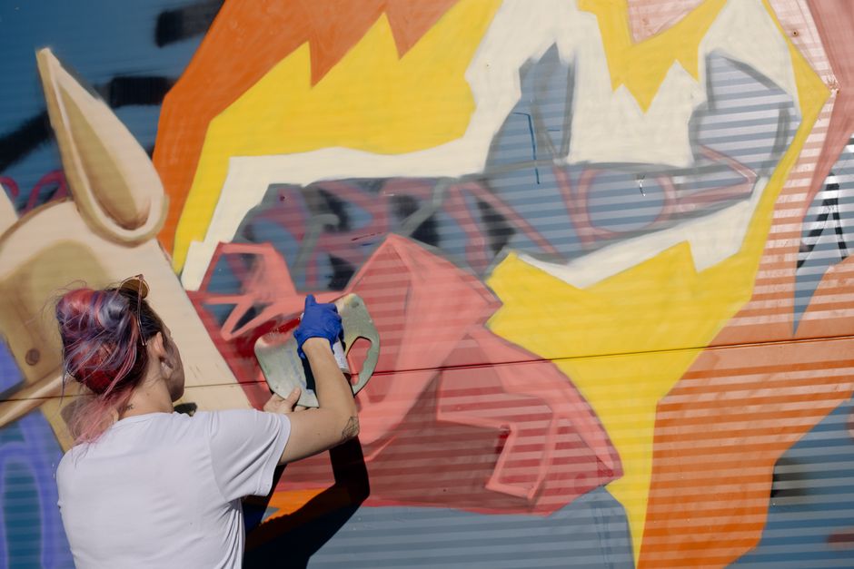 Female street artist spray painting a vibrant graffiti mural on a sunny day.